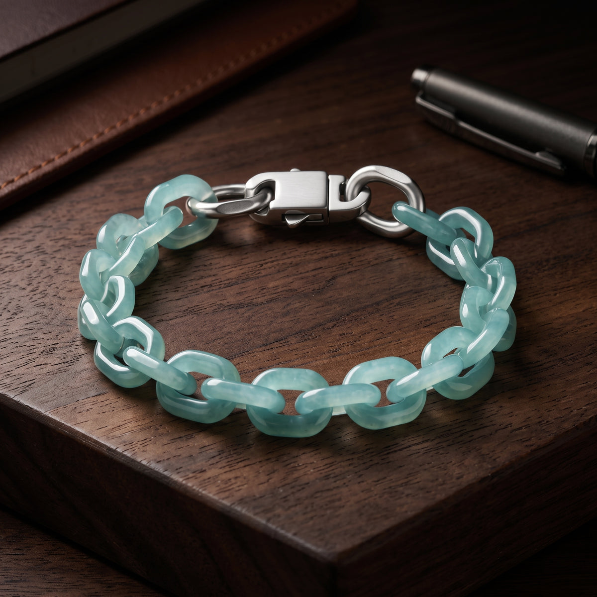 A bracelet made of translucent light aqua jade links connected by silver-toned metal rings and a silver clasp, displayed on a dark wooden surface with a pen and leather item in the background.