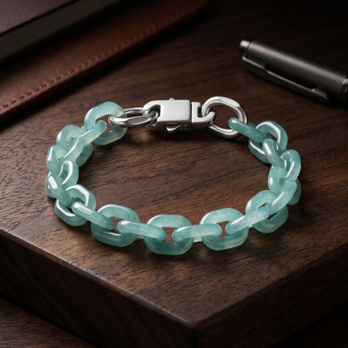 A bracelet made of translucent light aqua jade links connected by silver-toned metal rings and a silver clasp, displayed on a dark wooden surface with a pen and leather item in the background.