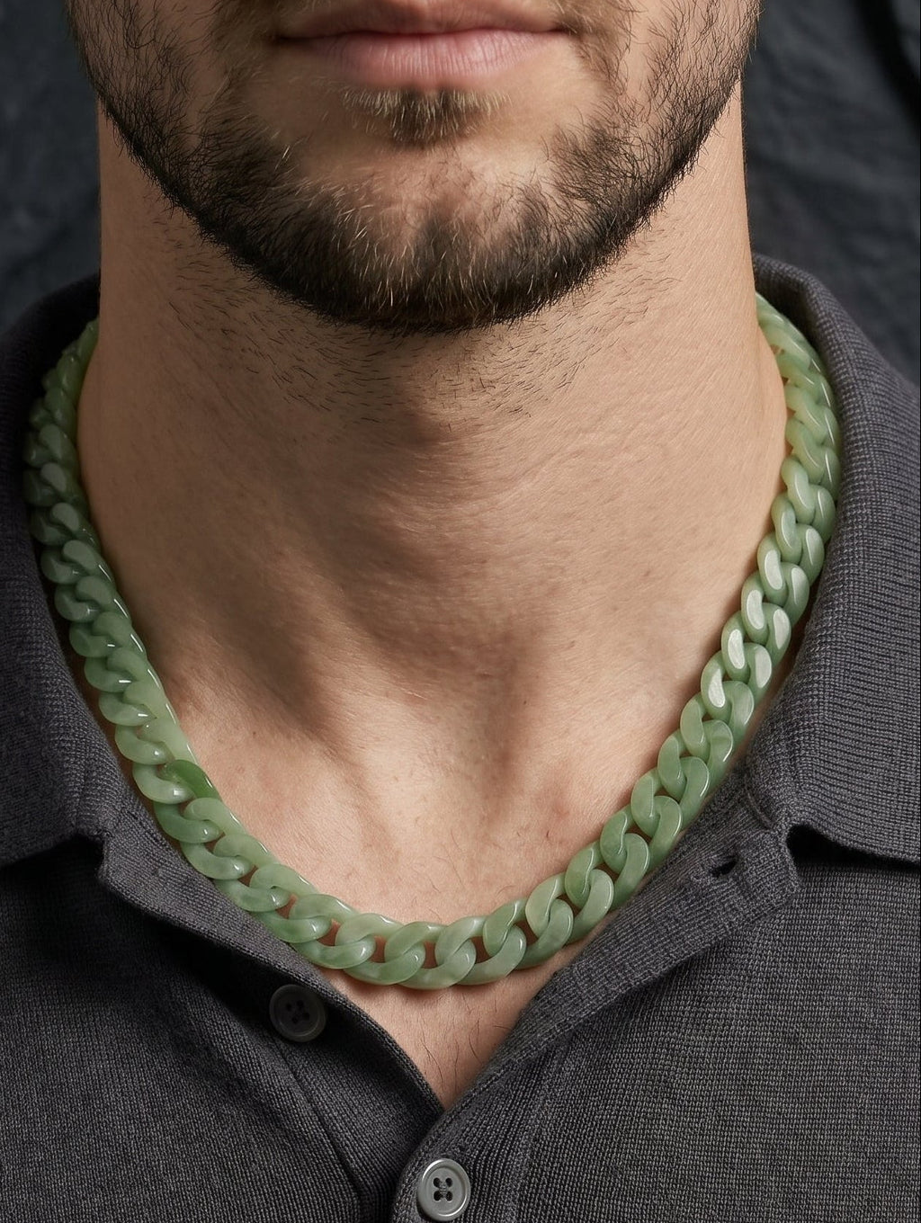 A close-up of a man wearing a chunky, light green jade chain-link necklace with a dark gray polo shirt, set against a dark textured background.