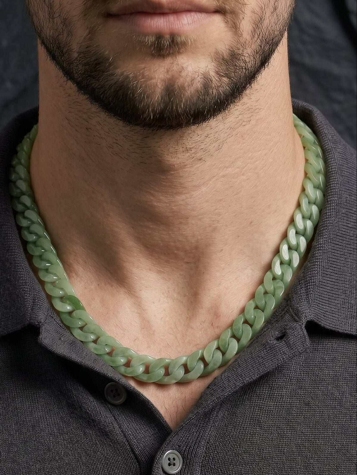 A close-up of a man wearing a chunky, light green jade chain-link necklace with a dark gray polo shirt, set against a dark textured background.