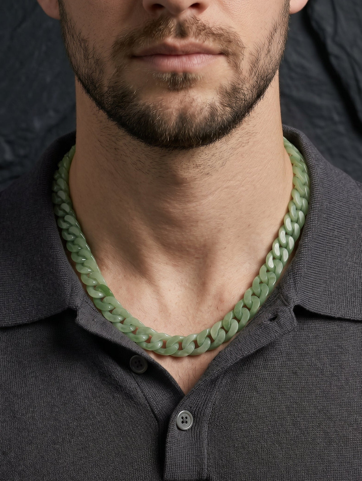 A close-up of a man wearing a chunky, light green jade chain-link necklace with a dark gray polo shirt, set against a dark textured background.